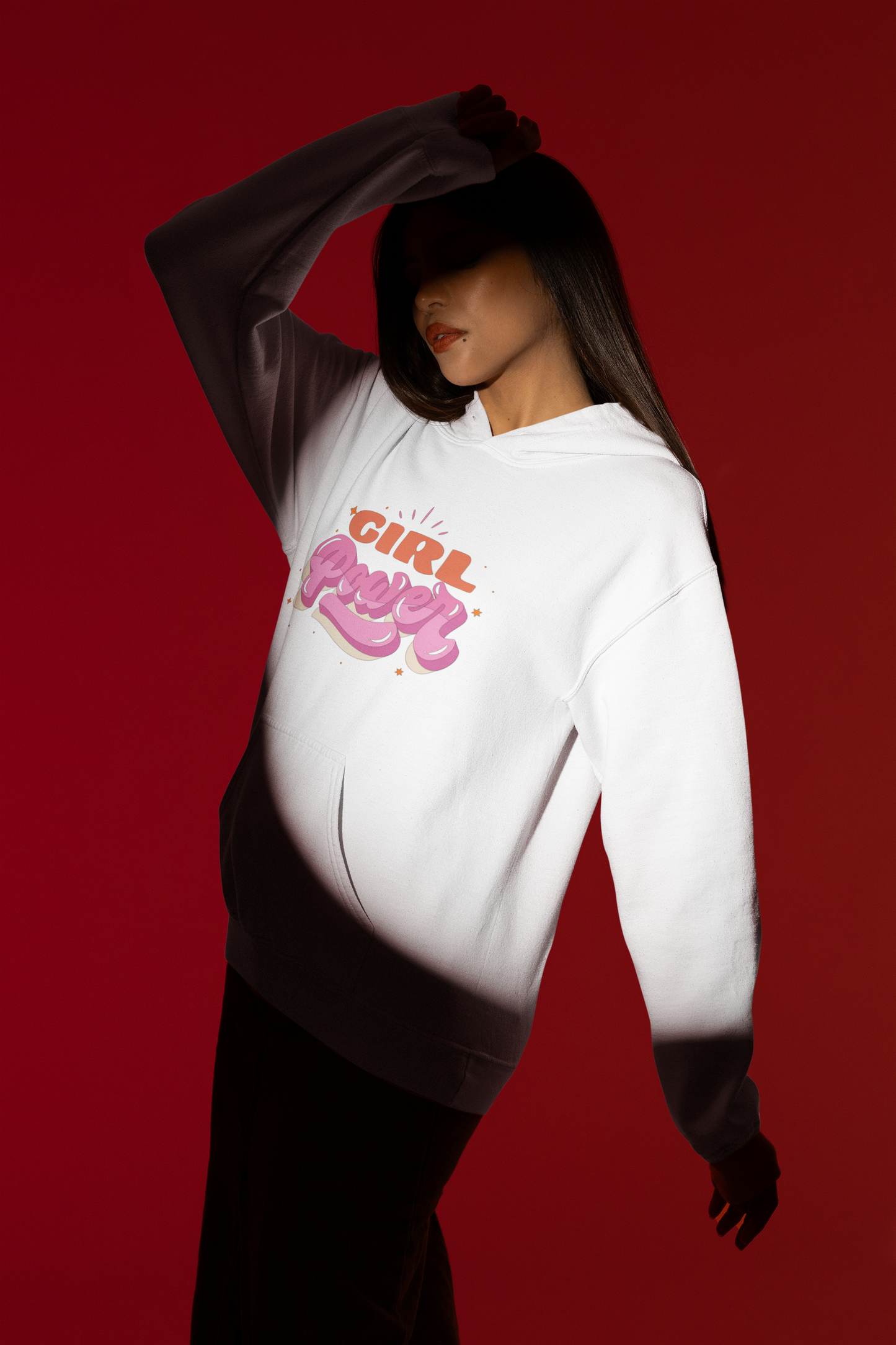 Wearing a Girl Power Hoodie, an individual stands against a red backdrop. Their face is partly shadowed, framed by flowing dark hair. With their right arm raised and partially concealing their head, they exude empowerment and the essence of women's rights gatherings.