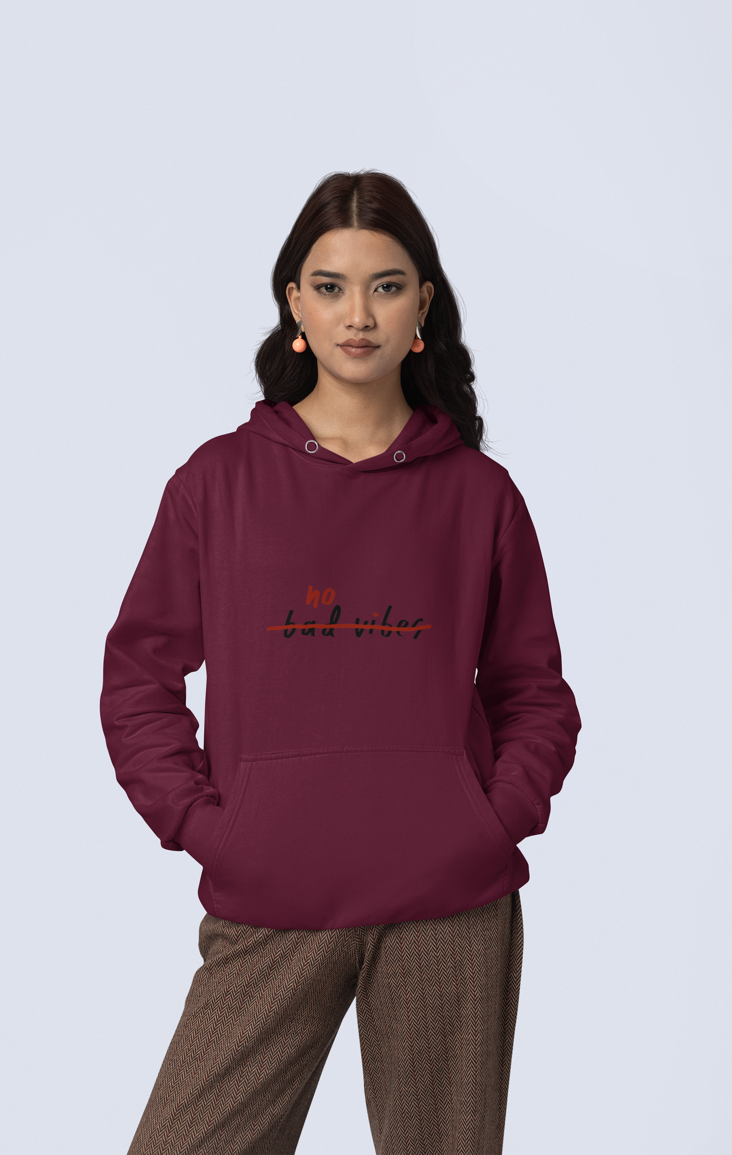 A person with long dark hair is set against a light gray background, dressed in a cozy and stylish hoodie called the No Bad Vibes Hoodie. This hoodie is produced from sustainable materials, and they pair it with brown patterned pants. Their hands are tucked comfortably into the front pocket of the hoodie.