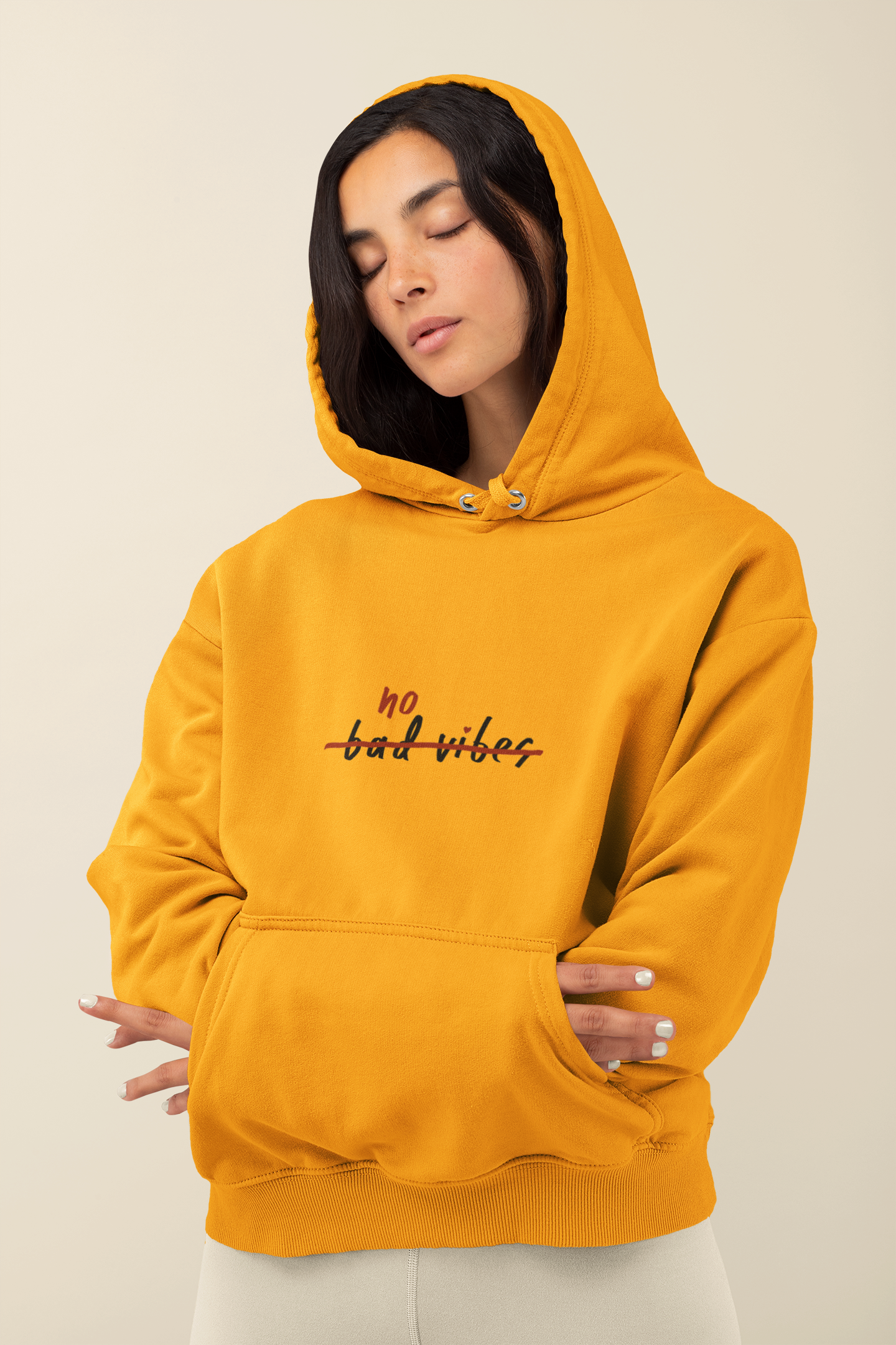 A person with long dark hair showcases a bright yellow No Bad Vibes Hoodie, known for its cozy and stylish design. Posed against a neutral backdrop, they have their eyes closed with hands nestled in the front pocket, emphasizing its sustainable production values.
