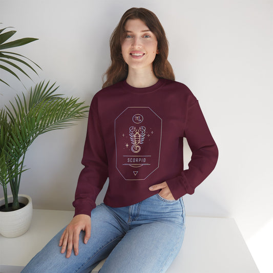 A woman with long brown hair, wearing a maroon Scorpio Sweatshirt and light blue jeans, sits on a white table beside a potted plant, smiling at the camera—perfect astrology gift for any star sign lover.