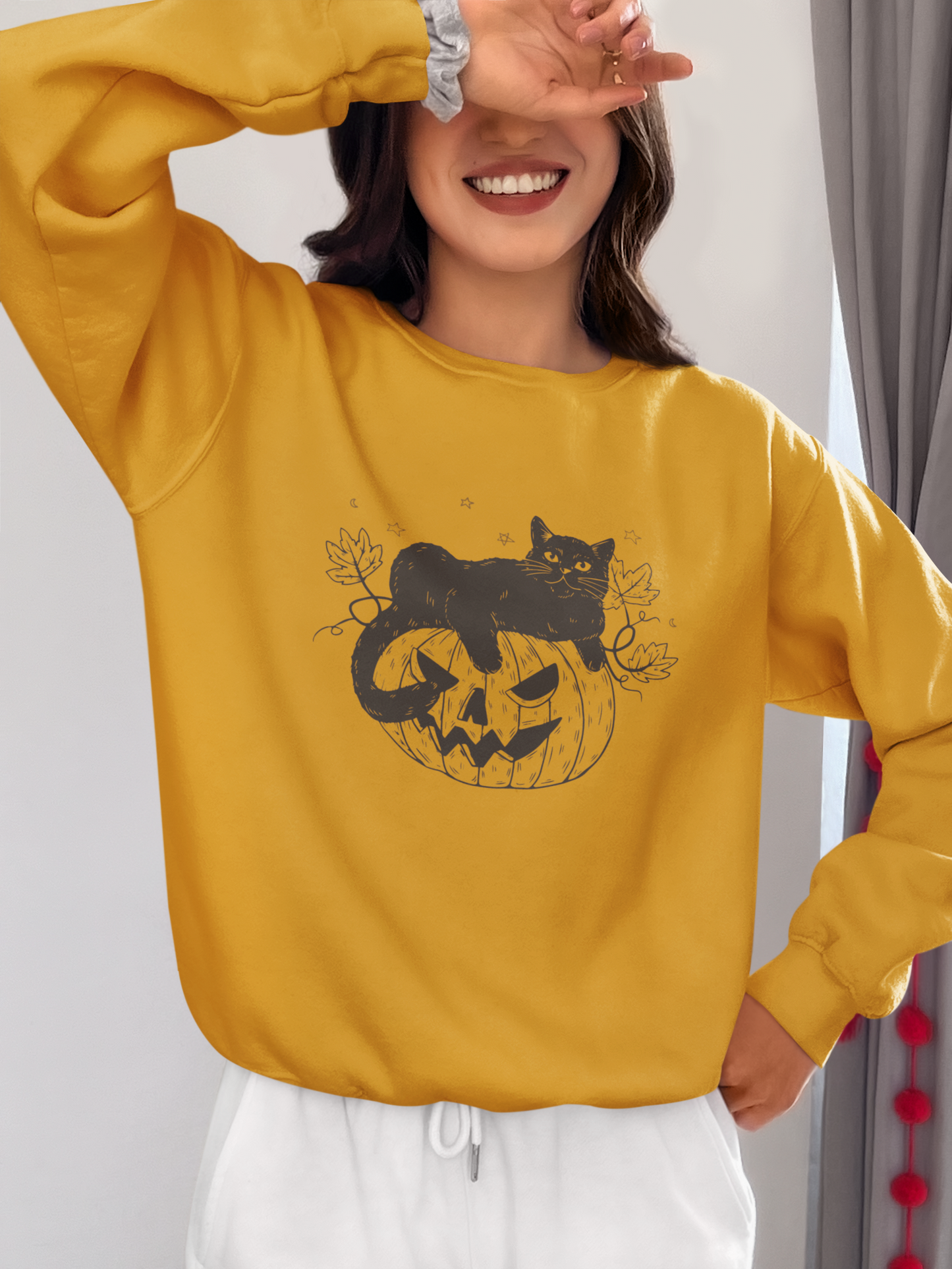 Wearing the Pumpkin Cat Sweatshirt—a yellow Halloween-themed garment perfect for cat lovers featuring a black cat sitting on a carved pumpkin—a woman playfully smiles while covering one eye with her hand, completing the spooky season look with white pants.