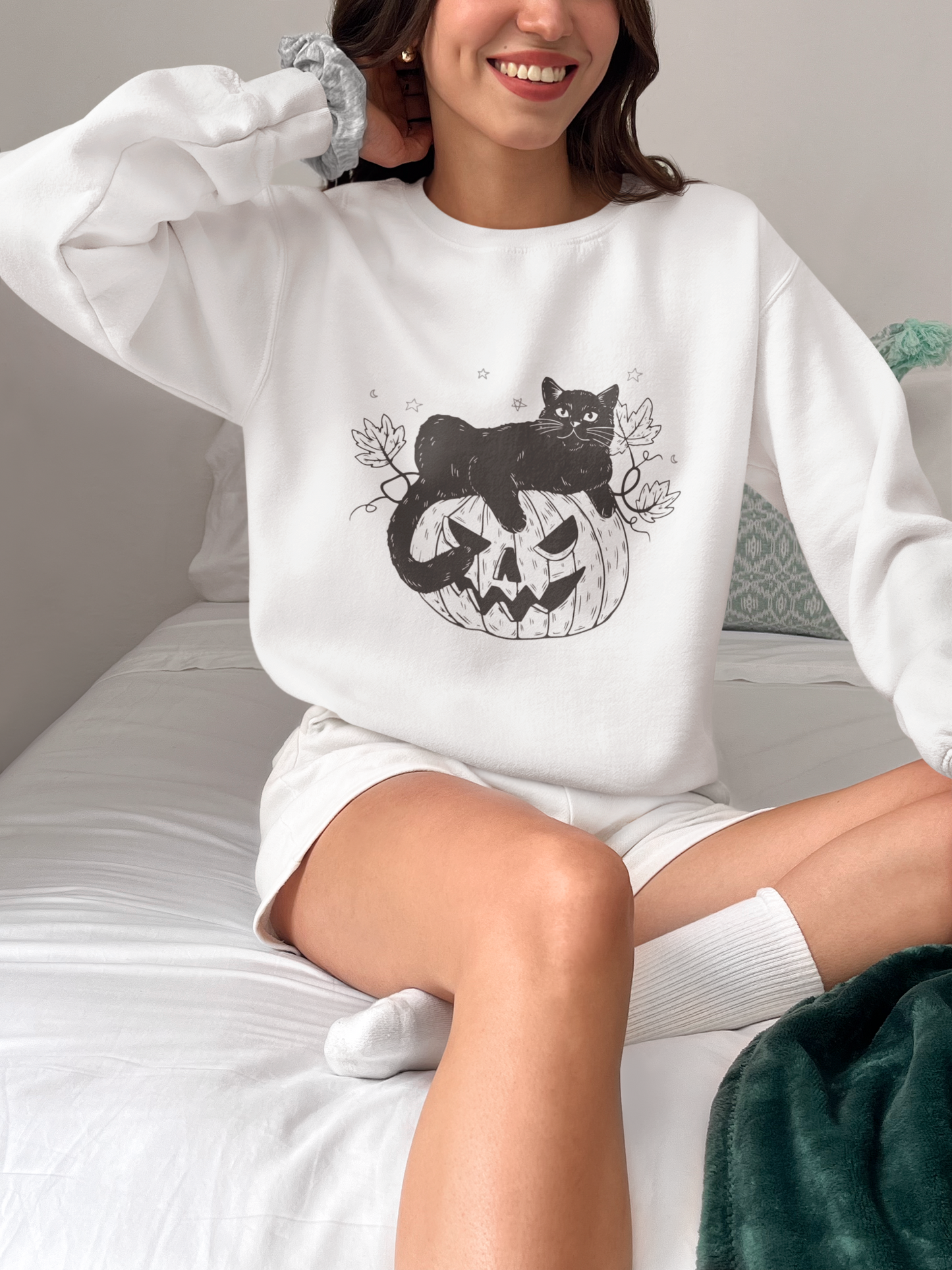 A person sitting on a bed, smiling, wearing the Pumpkin Cat Sweatshirt adorned with a black cat and pumpkin design. The room is filled with cozy, neutral tones, ideal for cat lovers celebrating the spooky season, with a green blanket neatly laid at the edge of the bed.